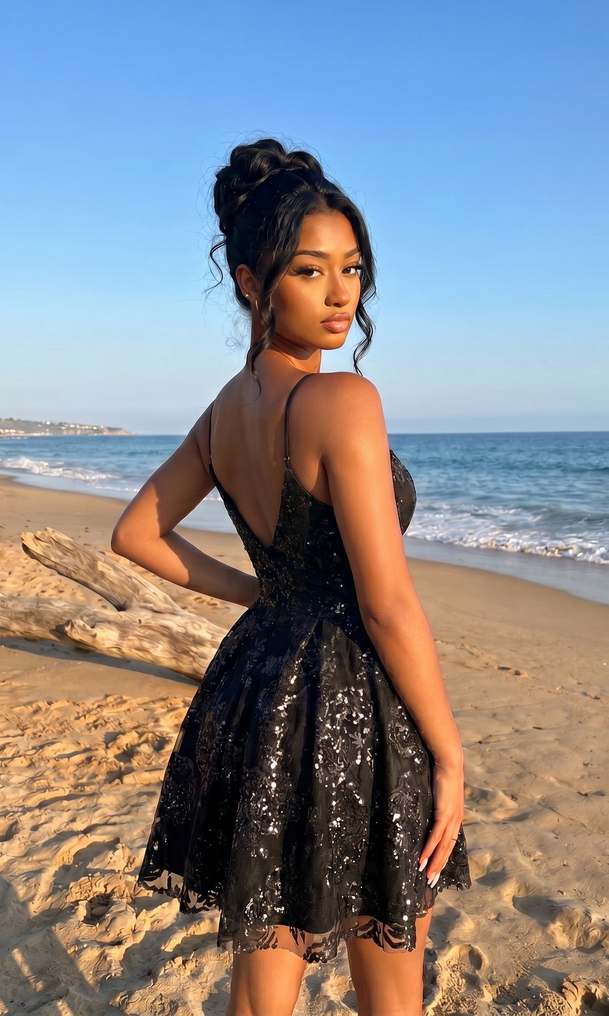 A woman in the Black Glitter Short Hoco Dress: Blondie Nites 5583BN stands on a sandy beach, facing the camera with the ocean and blue sky behind her.